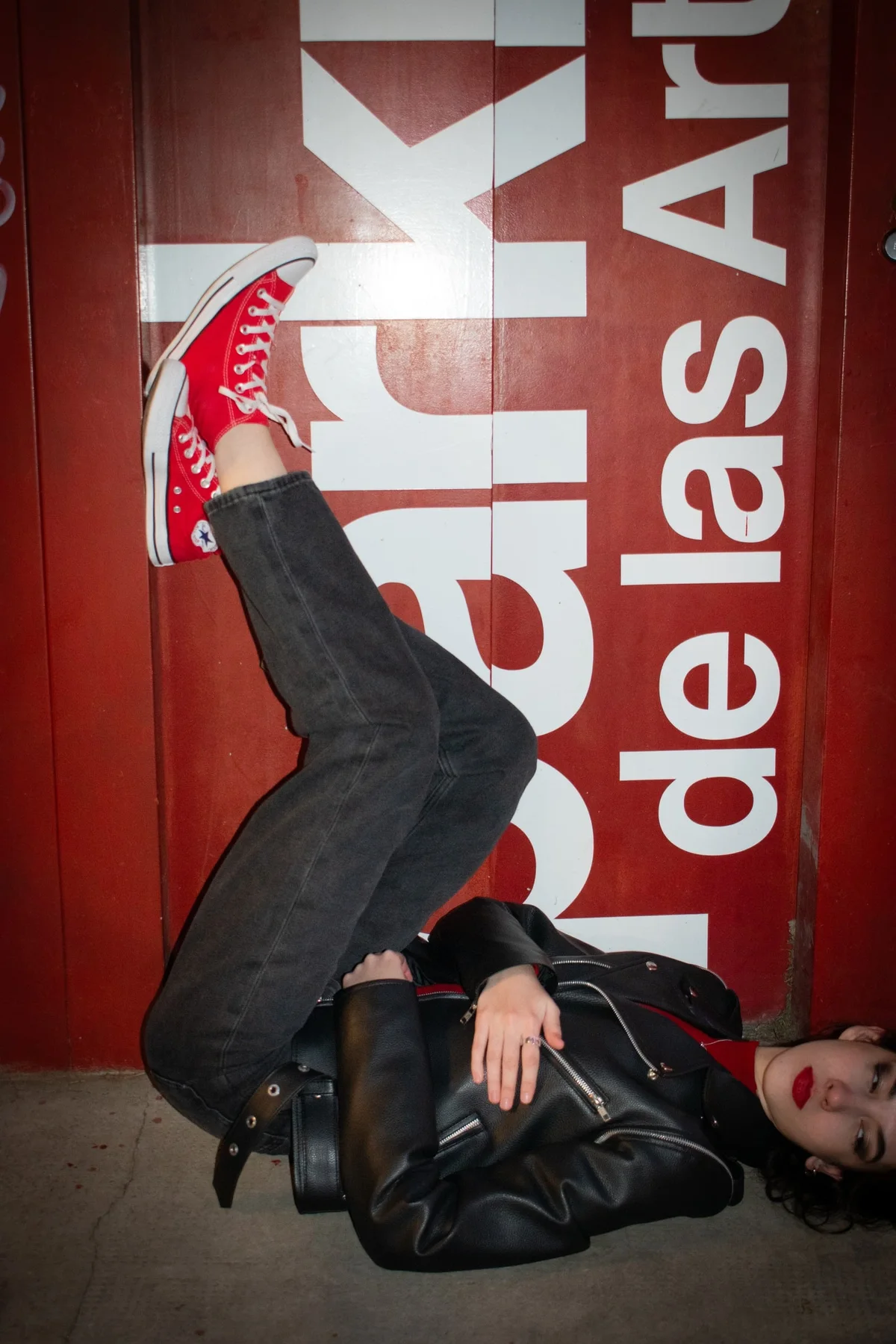 Modelo recostada en el piso con las piernas contra una pared roja y zapatillas Converse rojas