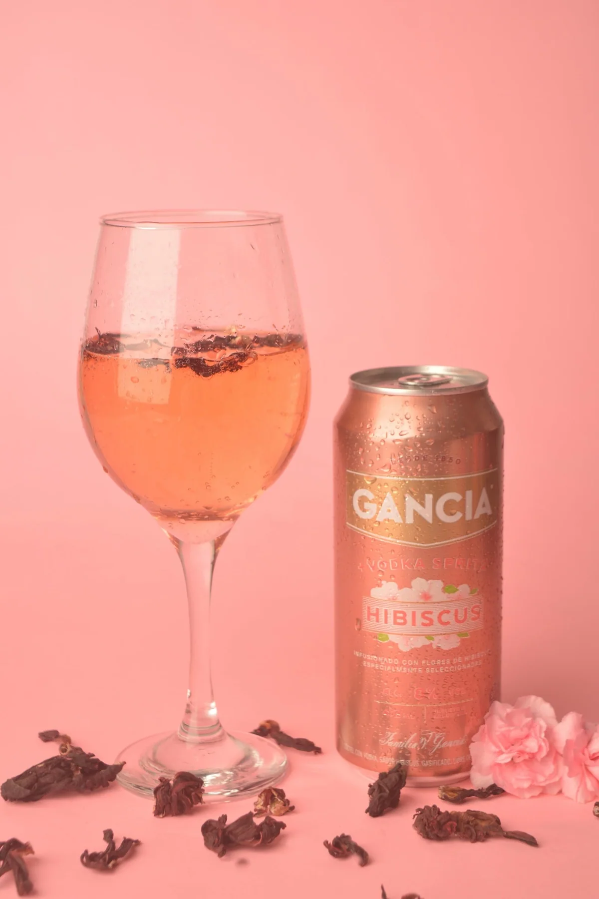 Copa de vino rosado con flores de hibisco junto a una lata de Gancia Hibiscus sobre fondo rosa