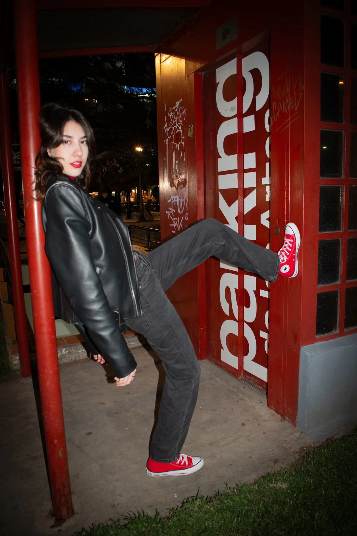 Modelo con campera de cuero patea con zapatillas Converse rojas junto a una puerta roja