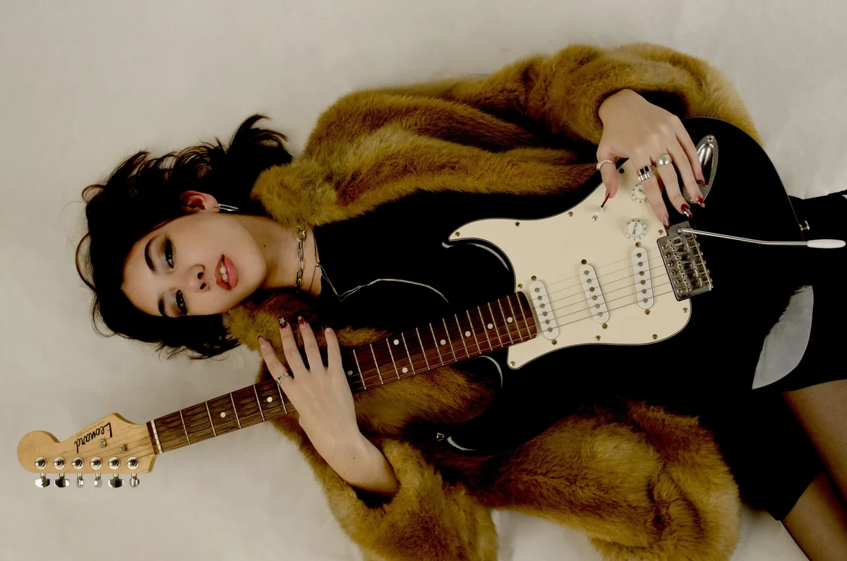 Modelo recostada en el piso con tapado de piel tocando una guitarra eléctrica